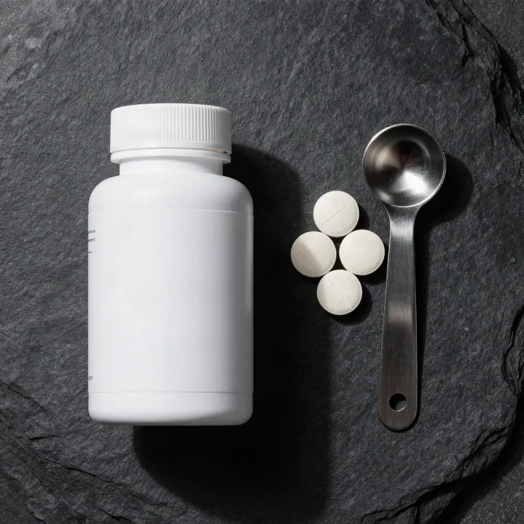 Overhead flat lay of a tablet bottle, four pale tablets and a scoop on charcoal stone.